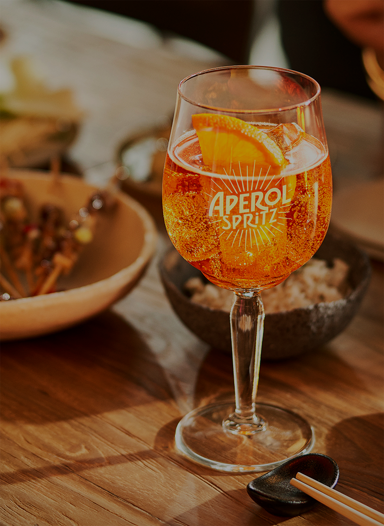 Aperol Spritz served in a branded glass with an orange slice, placed on a wooden table next to bowls of food and chopsticks.