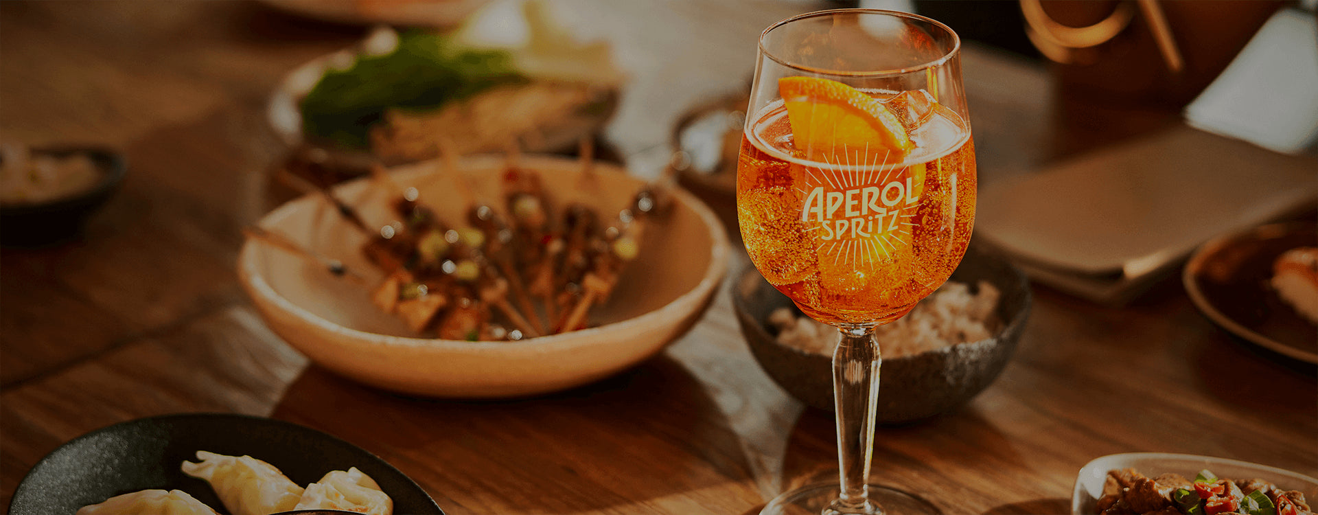 Close-up of an Aperol Spritz in a branded glass with an orange slice, surrounded by dishes of food on a wooden table.