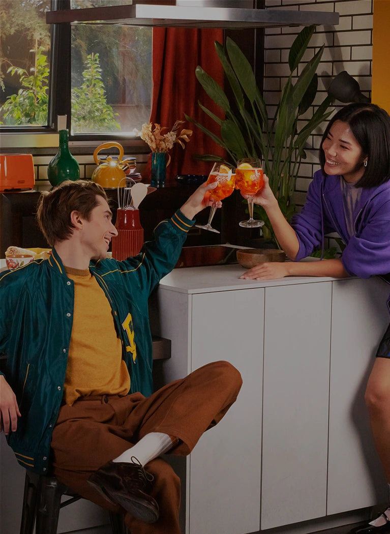 A man sitting and a woman leaning on a counter toast with Aperol Spritz glasses, smiling at each other in a cozy modern kitchen.