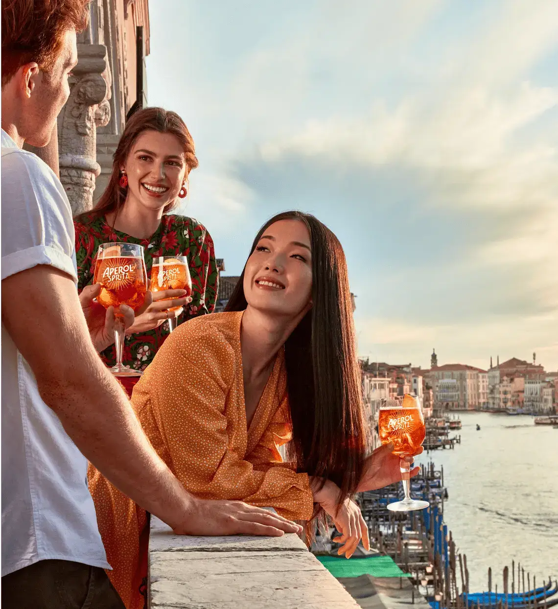 Three people enjoying Aperol Spritz on a balcony overlooking the Grand Canal in Venice at sunset.