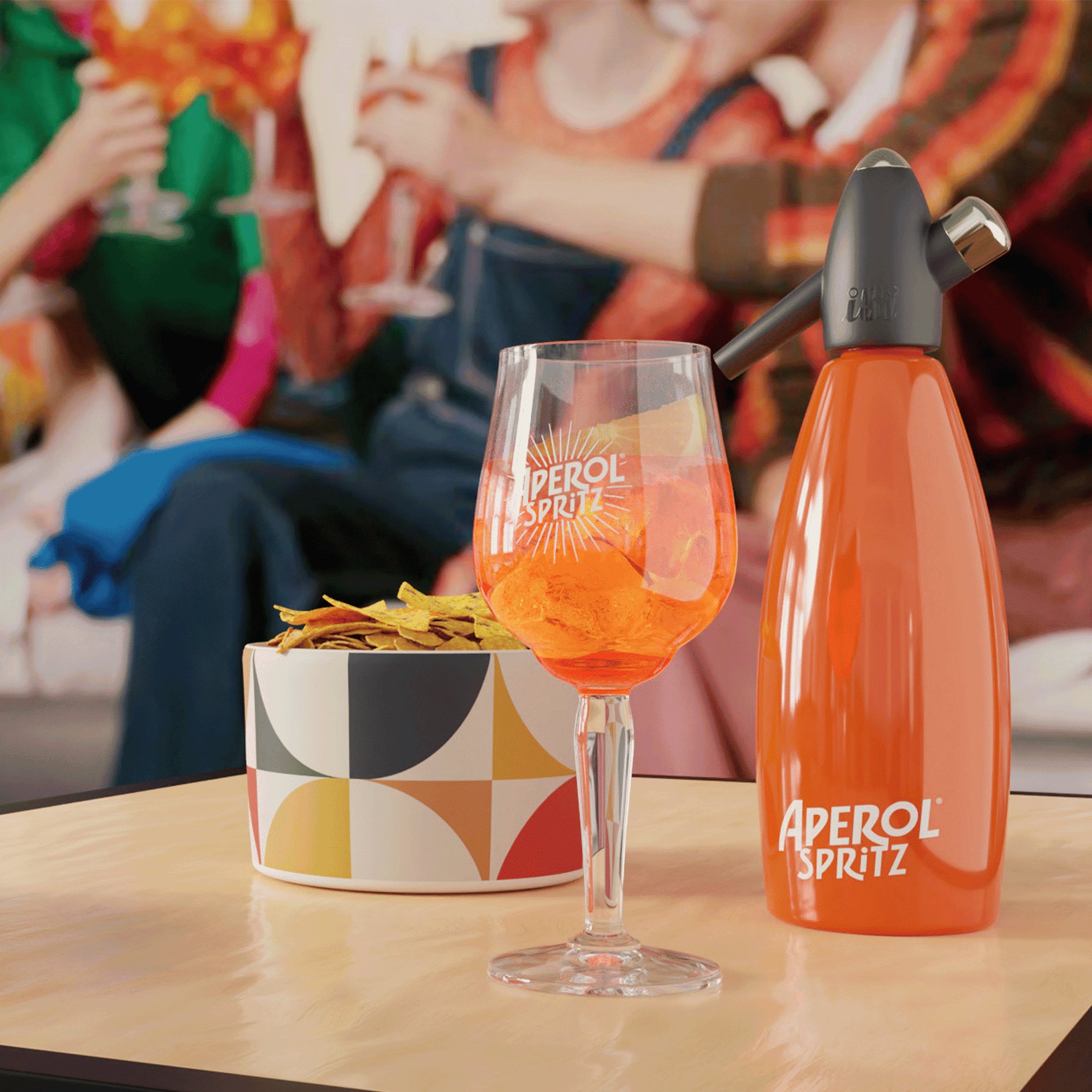 Aperol Spritz cocktail glass and orange siphon on a coffee table, next to a colorful bowl of chips, with a blurred background showing friends toasting.
