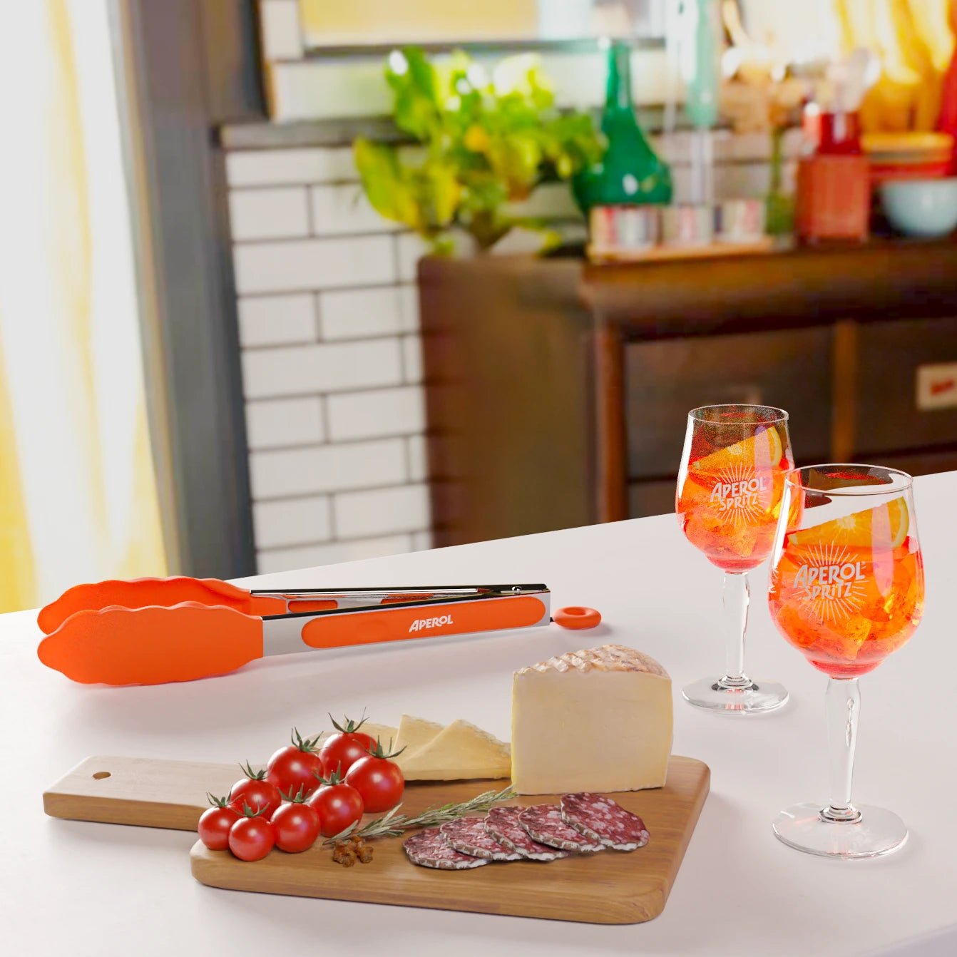 Two Aperol Spritz glasses on a white countertop next to a charcuterie board with cheese, cherry tomatoes, and salami slices, featuring an orange Aperol kitchen tong in the background, set in a cozy kitchen.