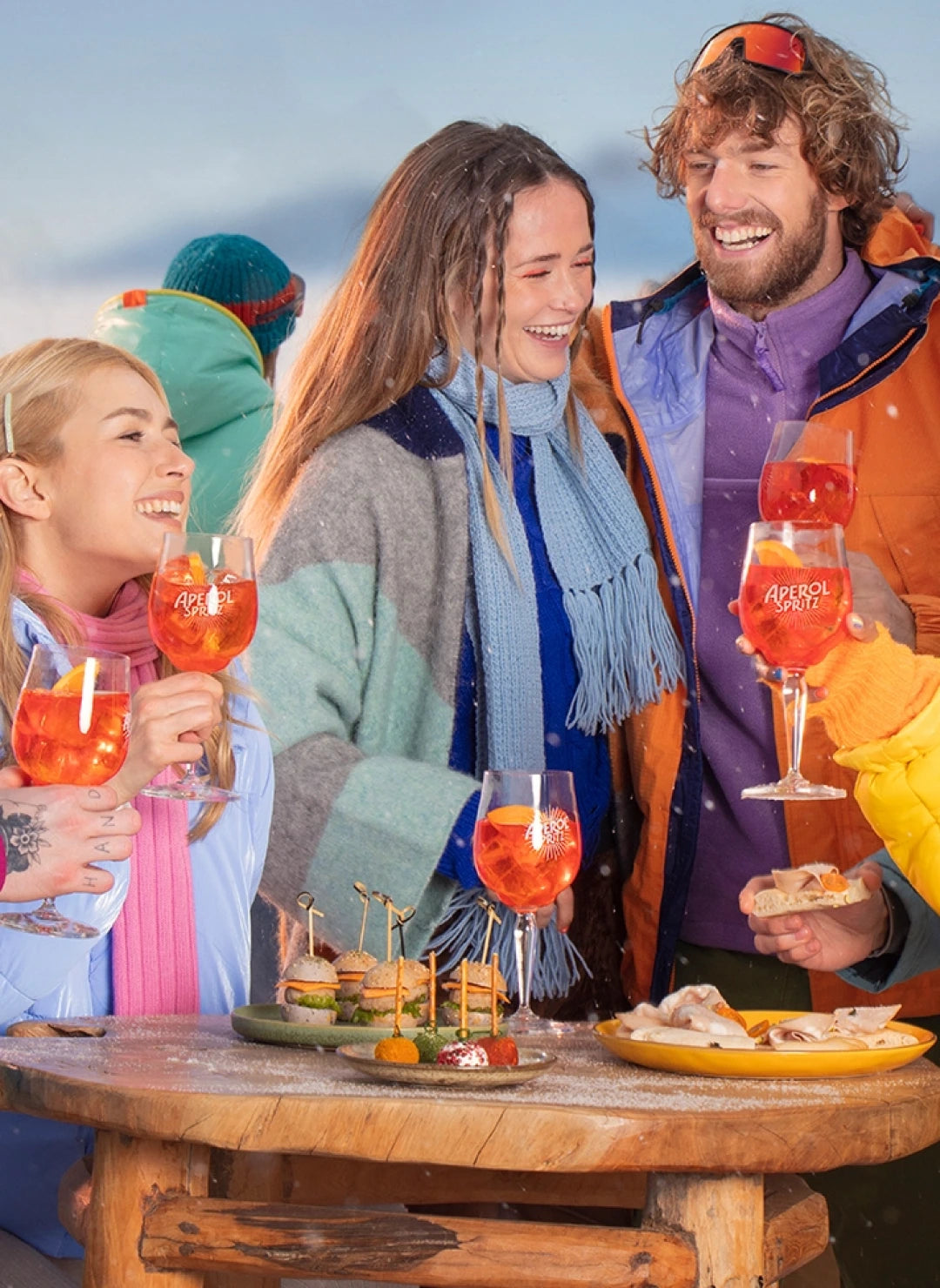 Smiling group enjoying Aperol Spritz and appetizers outdoors in the snow, dressed in colorful winter clothing