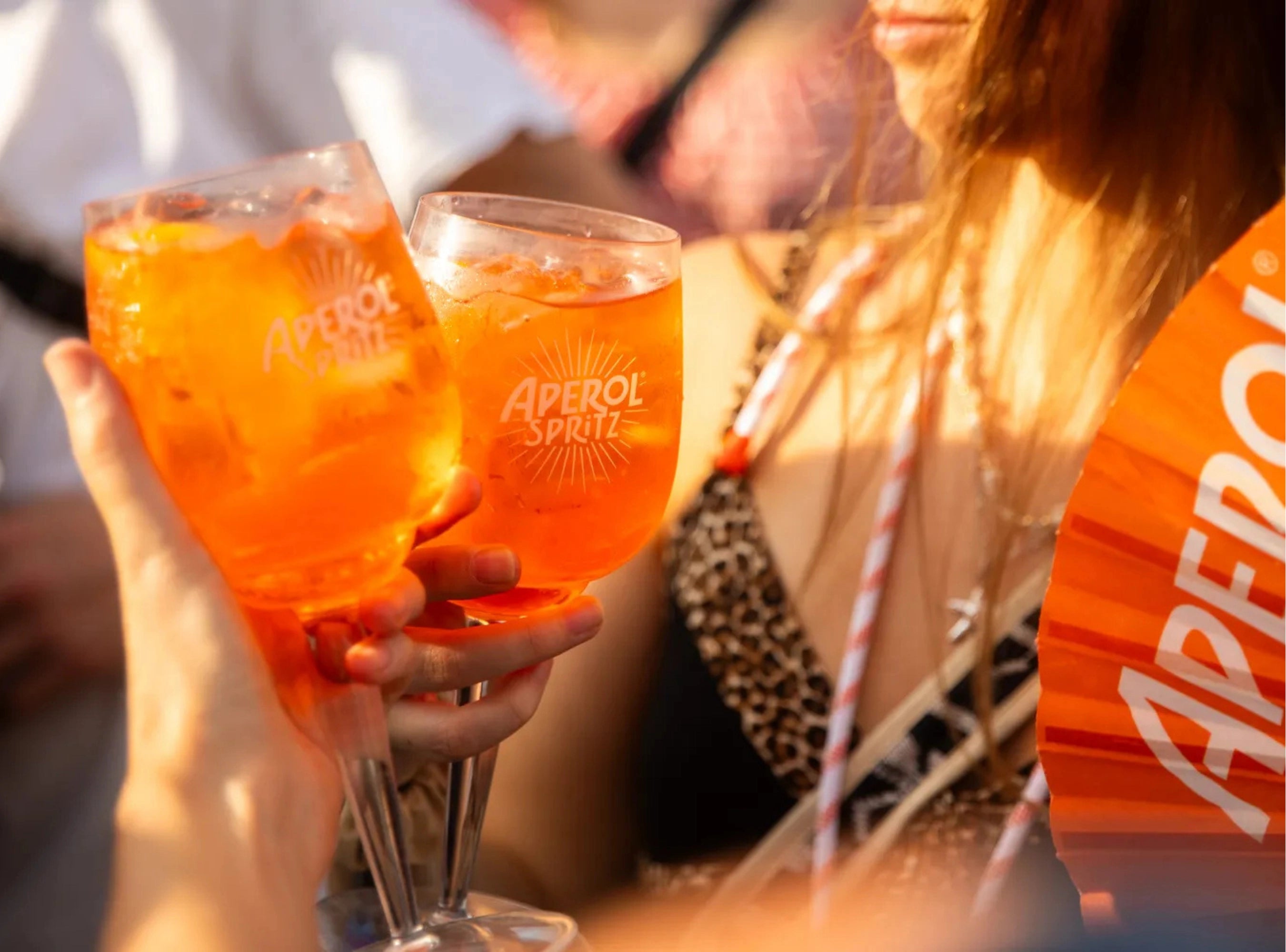 Two people clinking Aperol Spritz glasses filled with orange cocktail mix in warm sunlight, with an orange branded fan visible on the right.