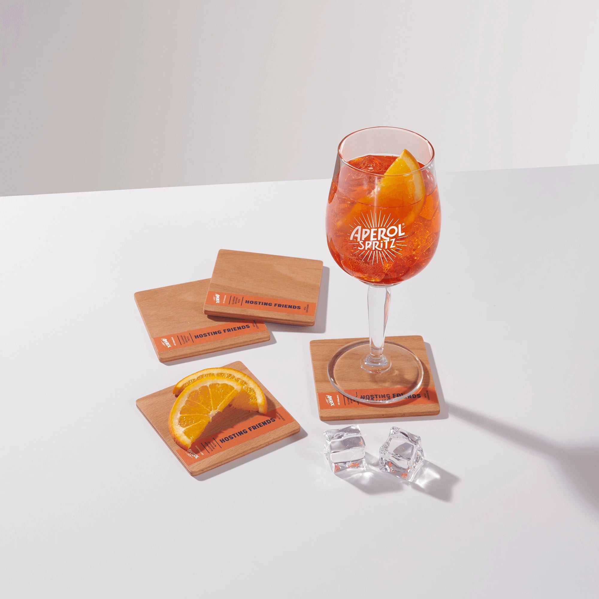 Aperol Spritz glass on a wooden coaster with orange slice garnish, surrounded by three additional coasters, ice cubes, and a coaster holding orange slices on a white surface.