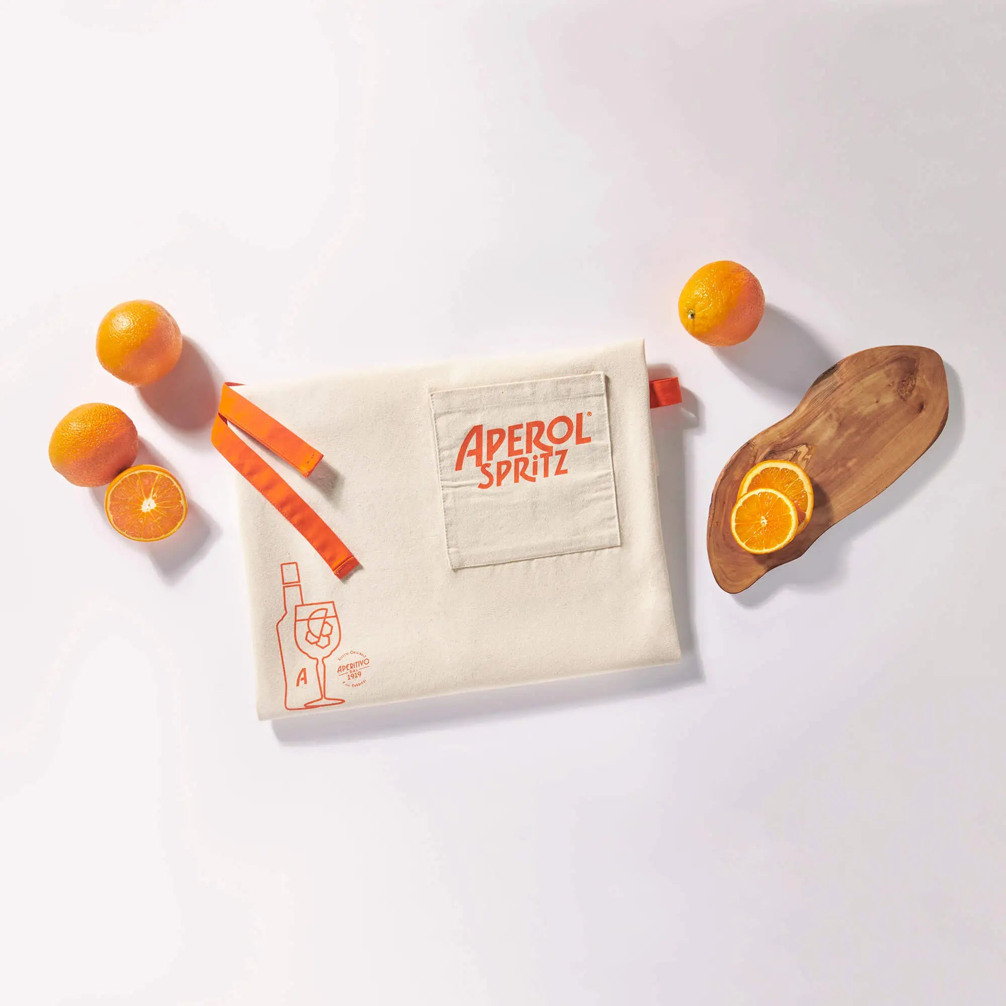 Folded Aperol Spritz apron with orange straps and logo on the front pocket, surrounded by fresh oranges and a wooden board with orange slices on a white surface.