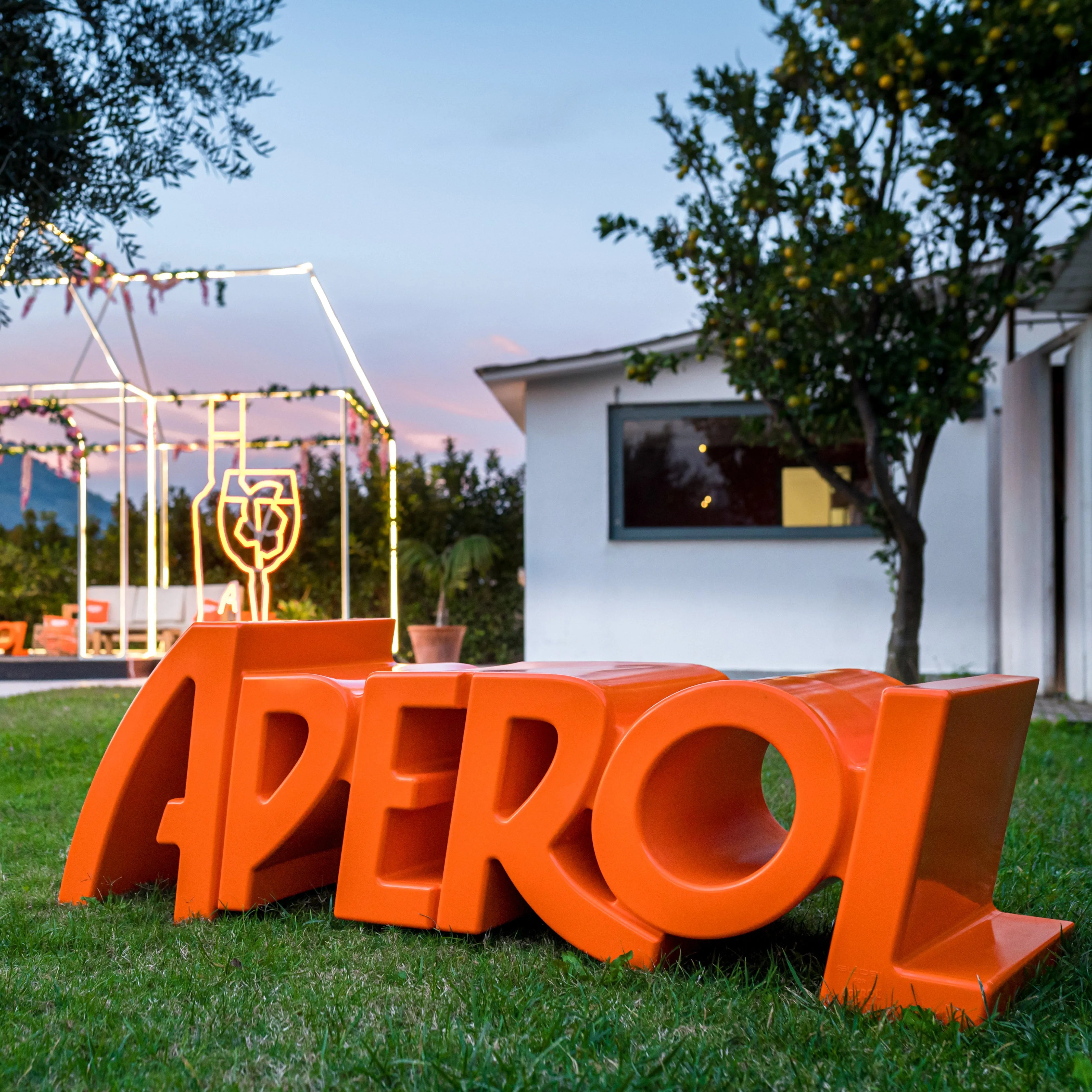 Outdoor Aperol-branded bench shaped like the bold orange letters of the word 'APEROL', placed on grass with a modern house and illuminated decorations in the background at sunset.