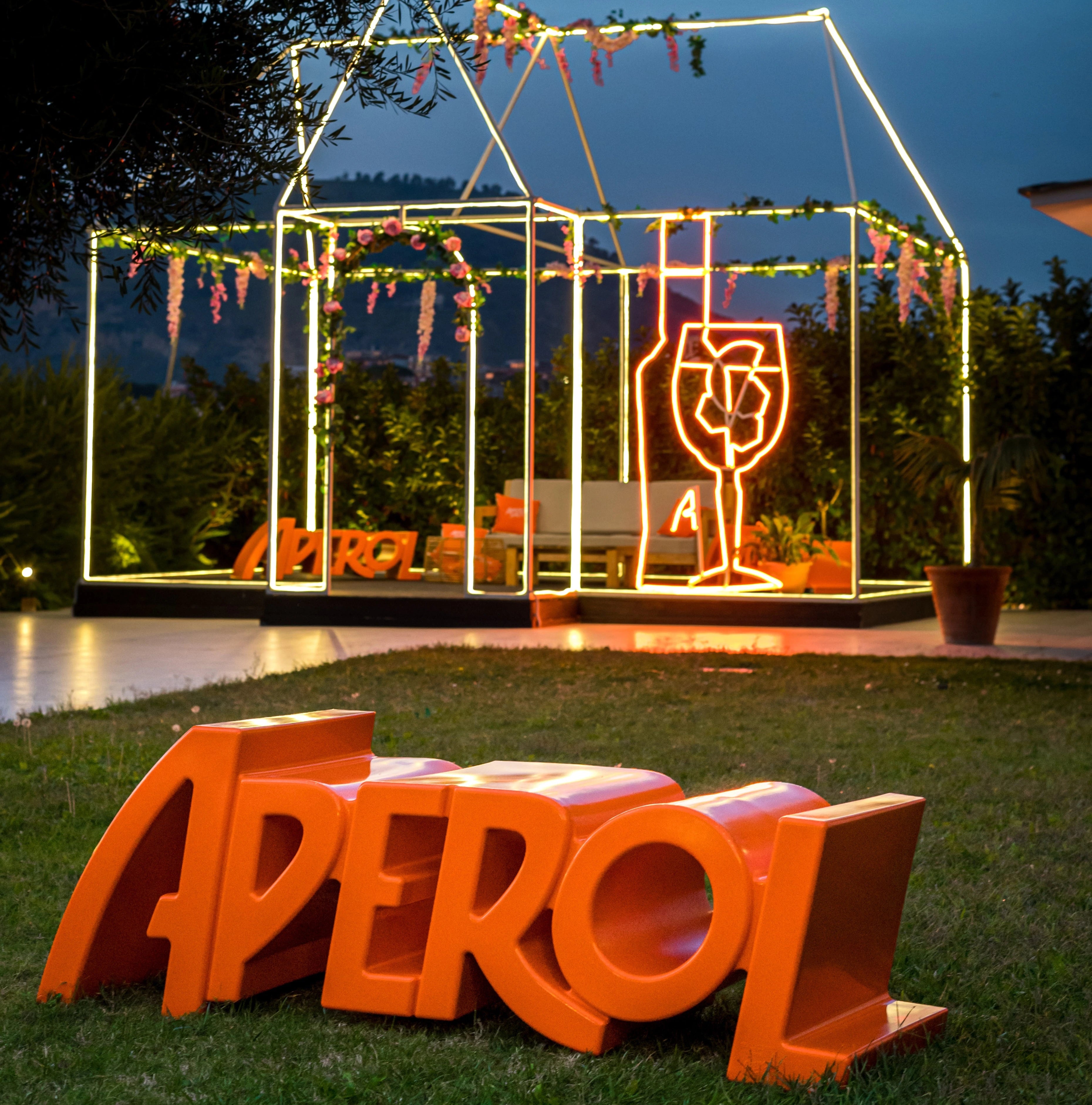 Orange Aperol bench in logo shape placed on lawn, with neon-lit outdoor lounge in the background