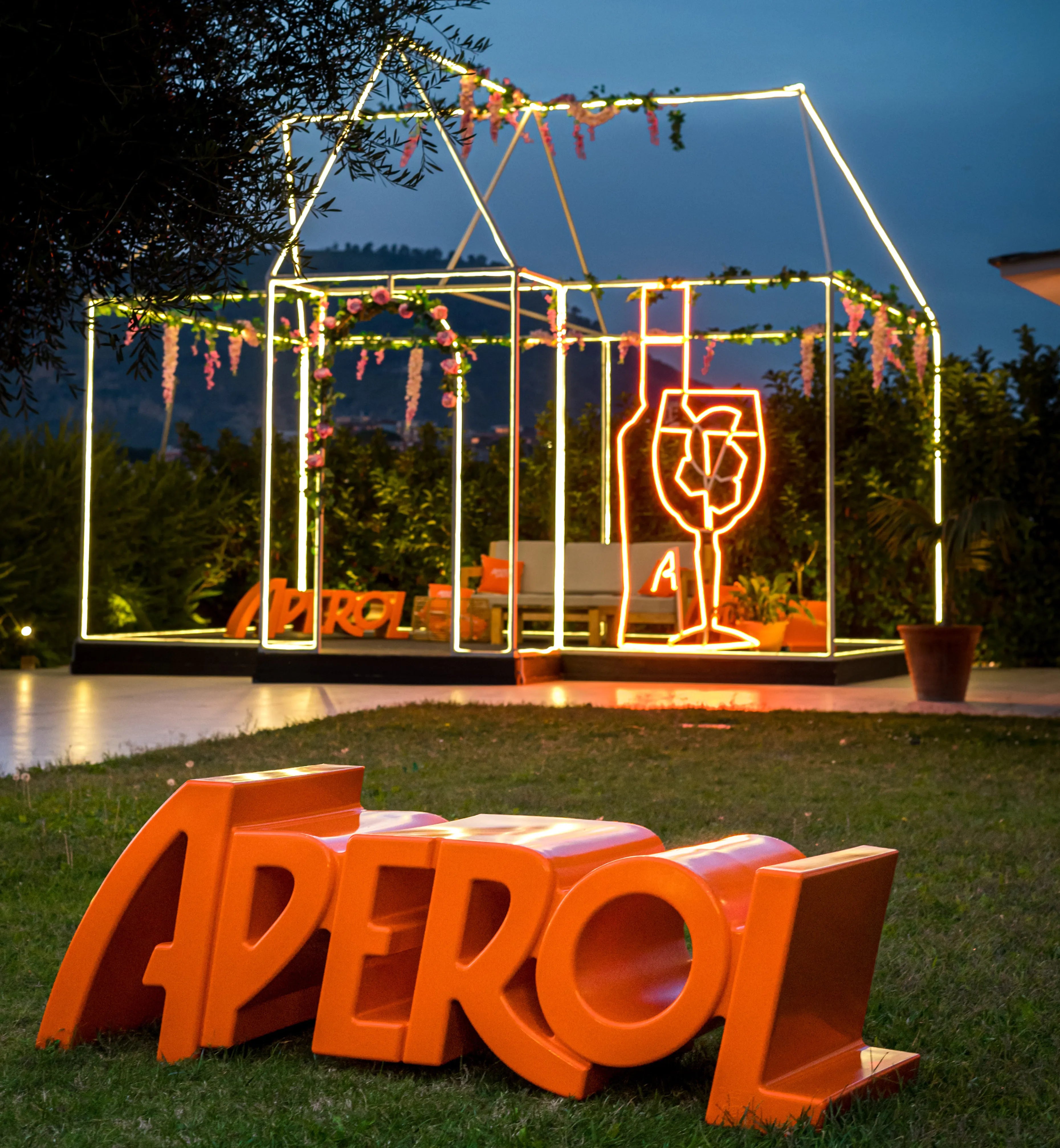 Orange Aperol-branded bench in the shape of the word 'APEROL' placed on grass, with a glowing neon garden structure and Aperol-themed decorations in the background during dusk.