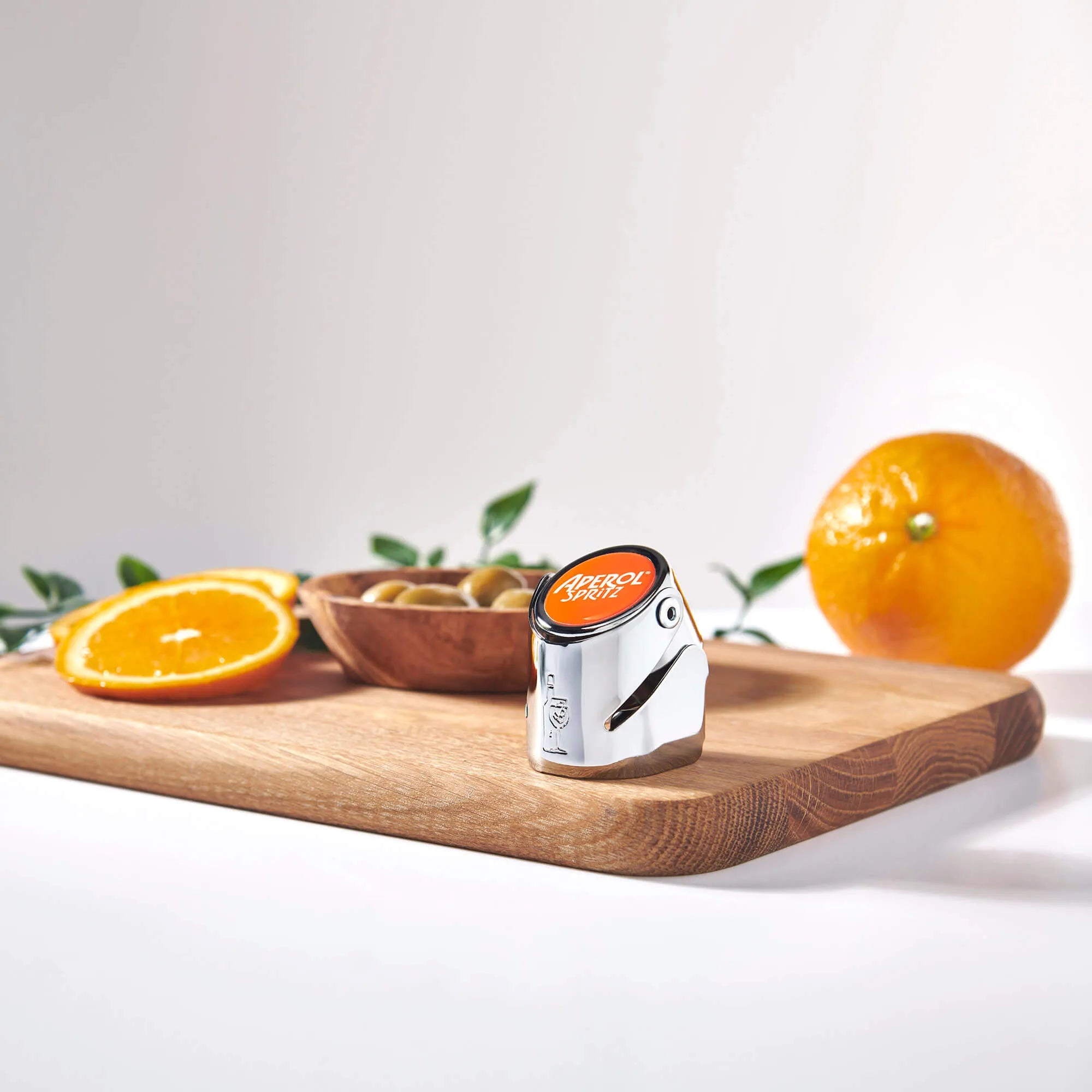 Aperol Spritz chrome bottle stopper with an orange top placed on a wooden cutting board, surrounded by sliced oranges, green olives in a wooden bowl, and a whole orange in the background.