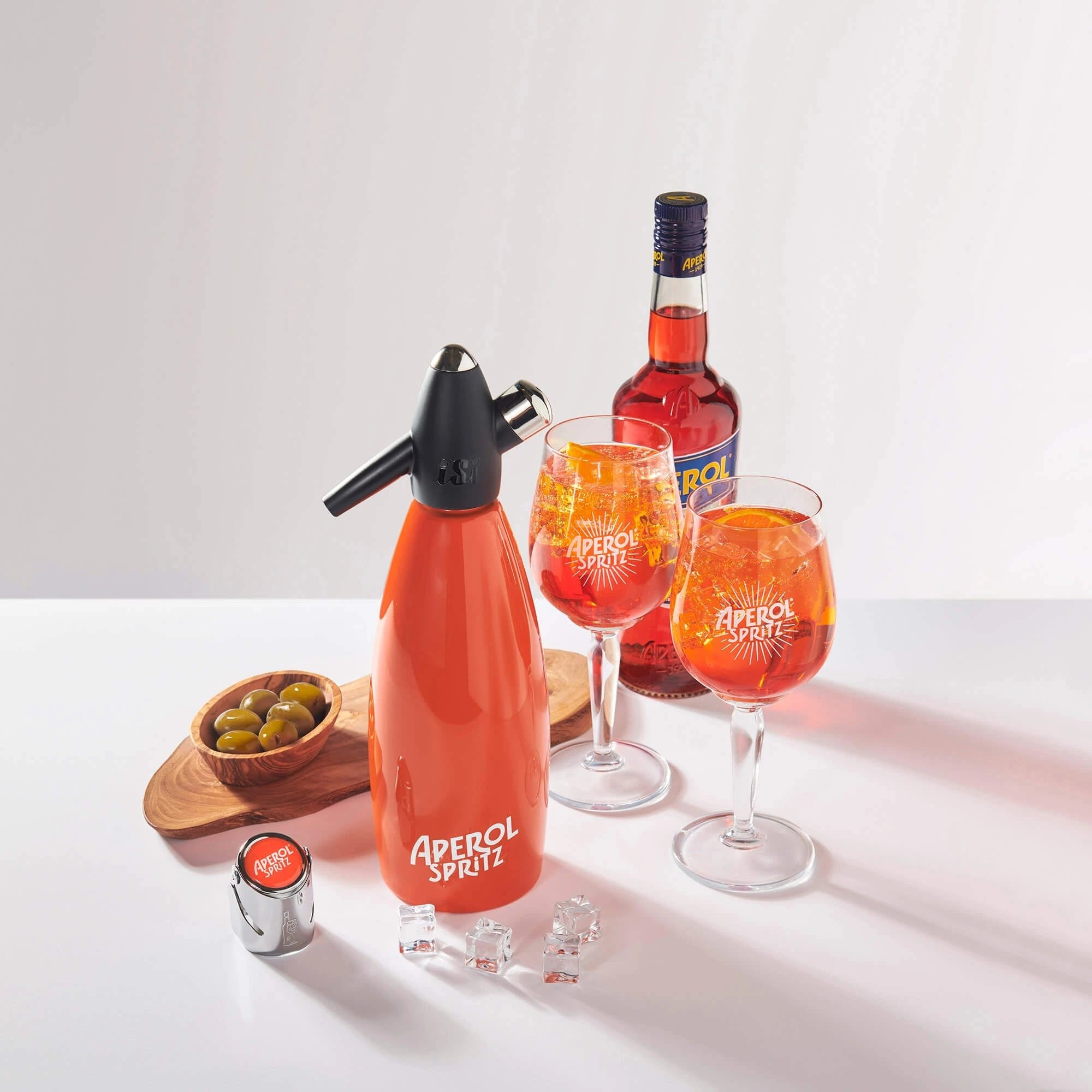 Aperol Spritz cocktail setup featuring a branded orange soda syphon, a bottle of Aperol, two Aperol Spritz glasses filled with the cocktail and orange slices, a champagne stopper, a wooden board with green olives, and a few ice cubes on a white table.