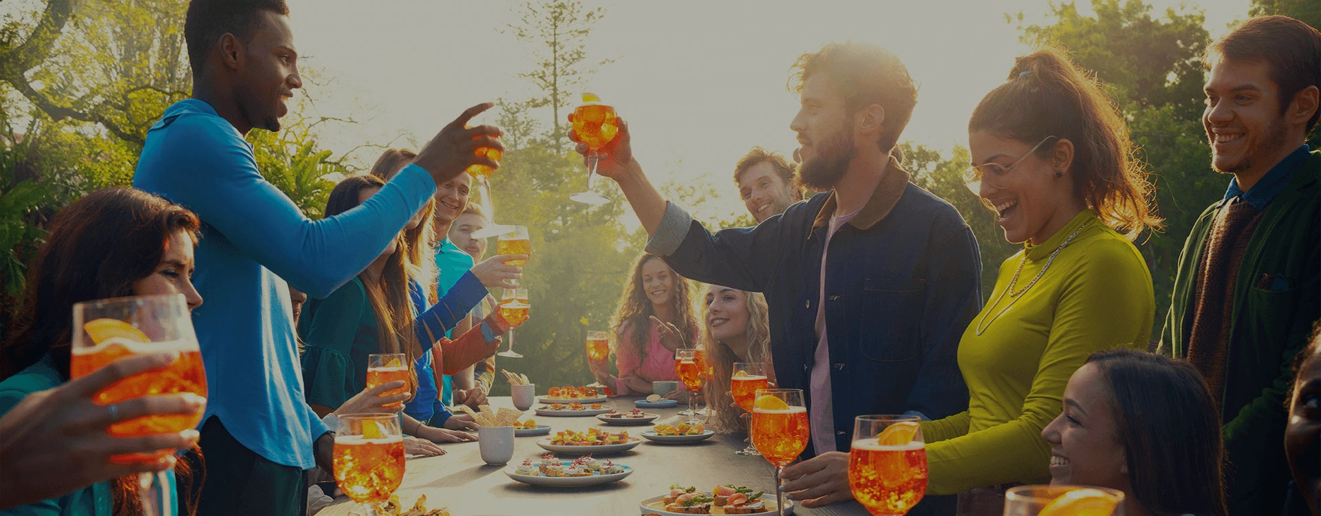 Group of friends enjoying Aperol Spritz cocktails outdoors, raising glasses together around a table filled with colorful appetizers under the sunlight.