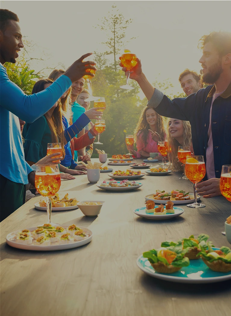 People gathered around a long table outdoors, toasting Aperol Spritz cocktails while sharing a variety of vibrant appetizers in a sunny setting.
