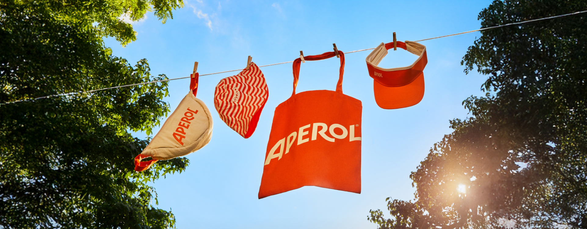 Aperol branded items including a bag, hat, and umbrella hanging on a clothesline with trees and blue sky in the background.