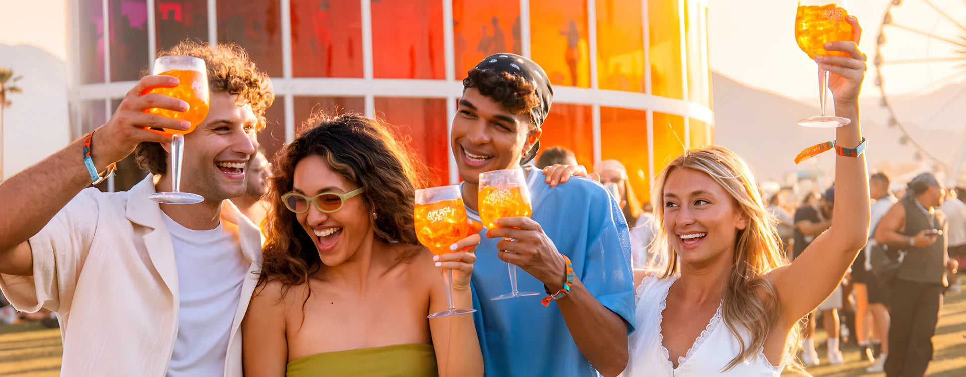 Group of four friends celebrating outdoors at a sunny summer event, raising Aperol Spritz glasses in a cheerful and festive atmosphere.