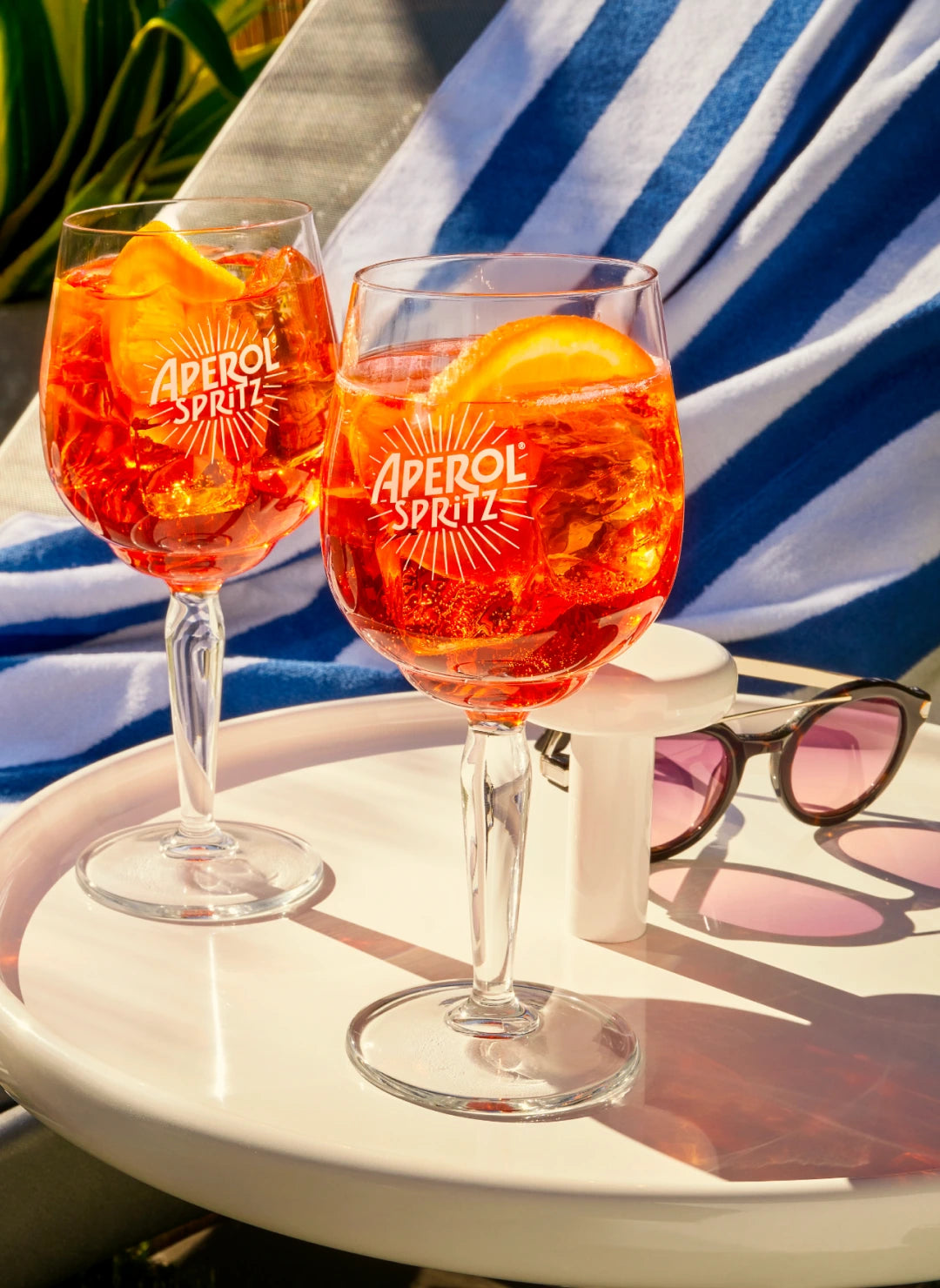 Two Aperol Spritz cocktails on a white table with sunglasses, beside a striped towel on a lounge chair