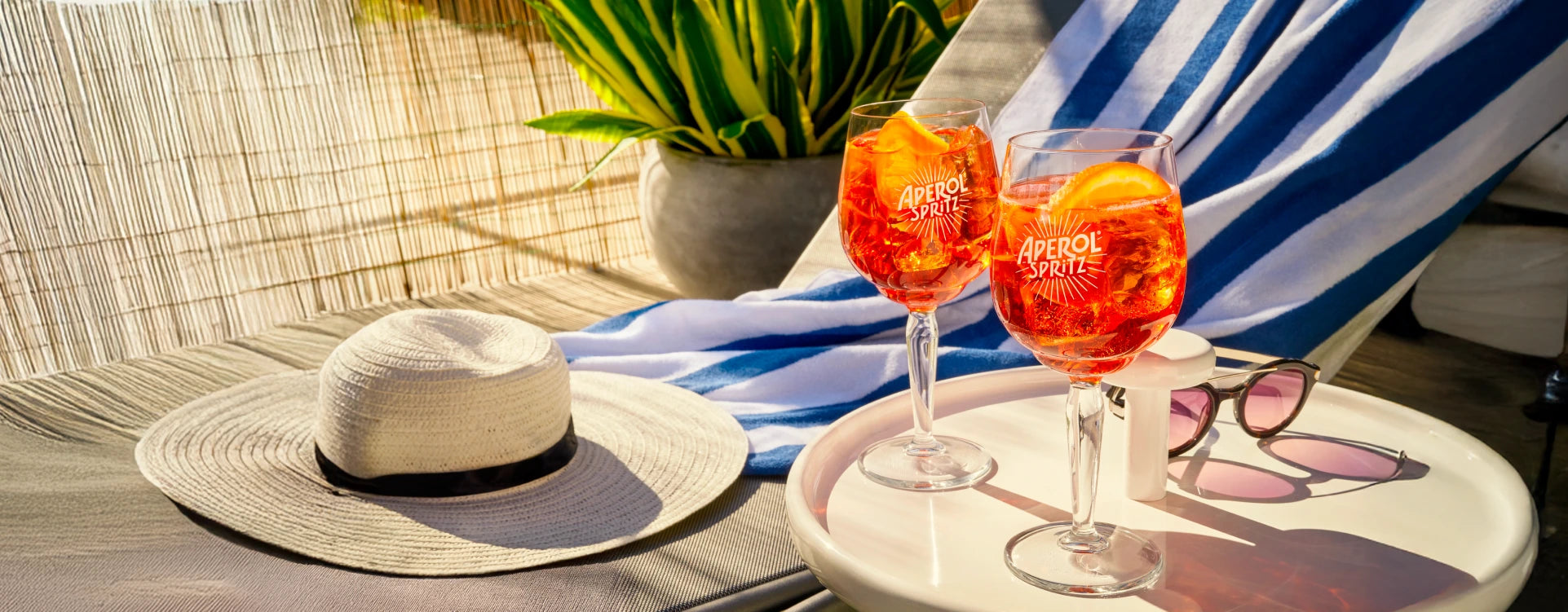 Two Aperol Spritz cocktails on a sunlit table with sunglasses, straw hat, and striped towel by a lounge chair