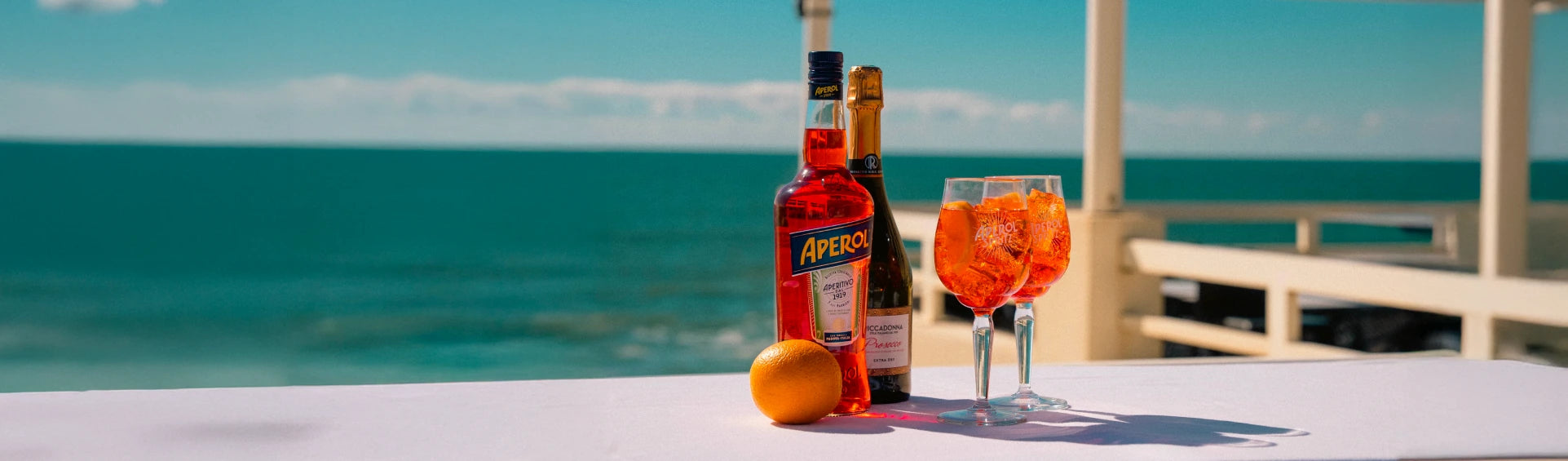 Aperol and sparkling wine bottles with orange and two Aperol Spritz cocktails on a table by the sea