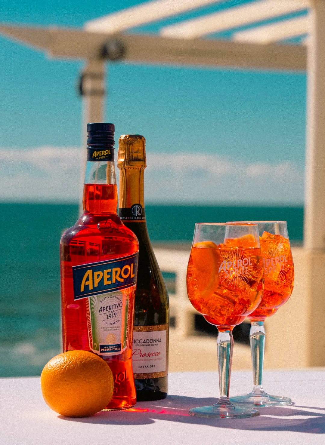 Aperol and prosecco bottles with orange and two Aperol Spritz cocktails on table by the sea under summer sun