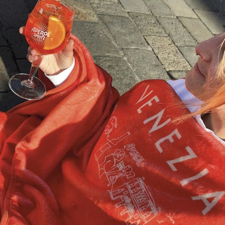Person wearing a red hoodie with 'Venezia' branding, holding an Aperol Spritz.